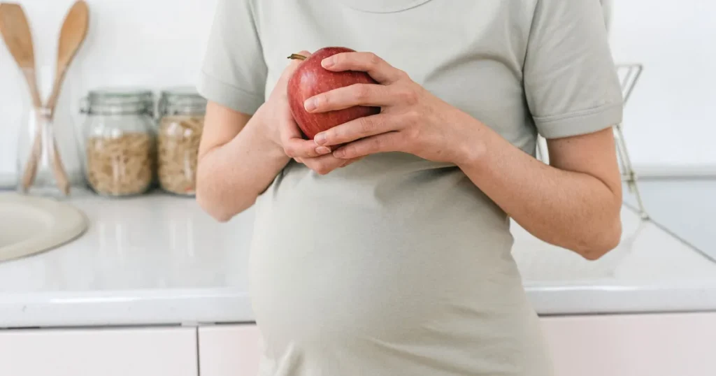 Gestante segurando uma maçã na cozinha, com a barriga em evidência