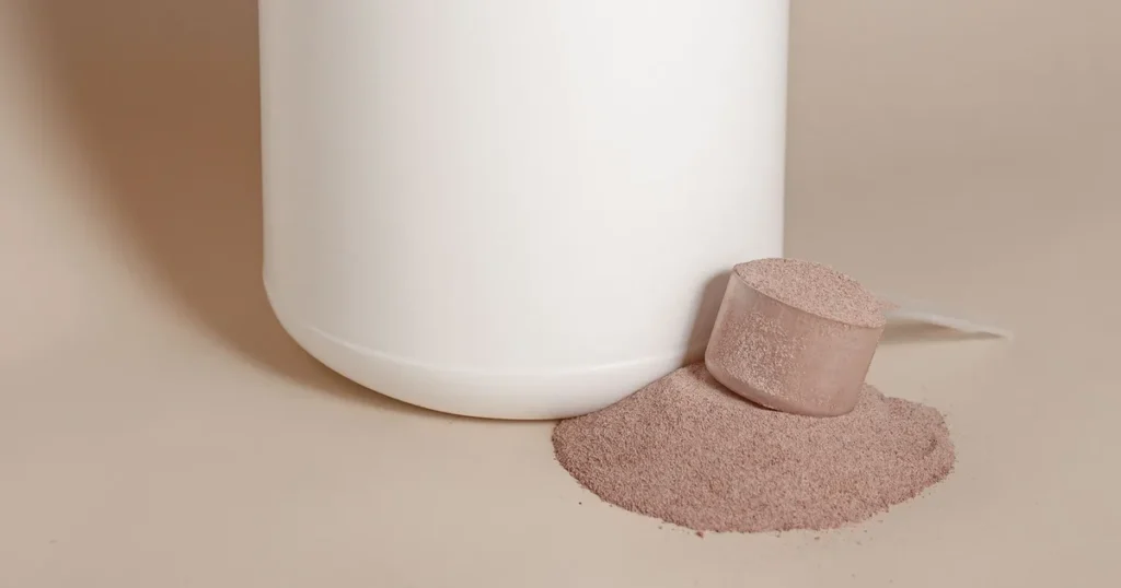 Scoop cheio de pó de whey protein ao lado do pote, com pó derramado na mesa
