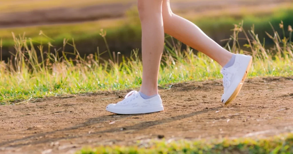 Pernas de uma pessoa caminhando de tênis branco num caminho de terra ao sol