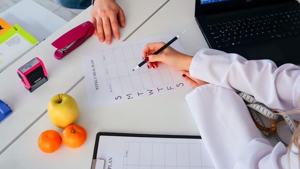 Nutricionista preenchendo um plano alimentar semanal durante a consulta, com frutas e fita métrica sobre a mesa